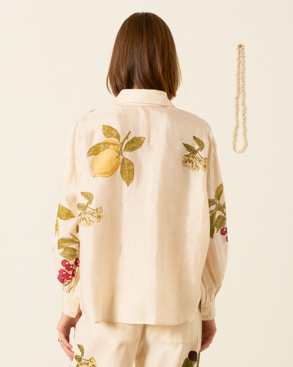 A person stands facing away, wearing the Emery Four Fruit Silk Shirt—a cream-colored, artisan piece with hand-block-printed fruit and leaf motifs. A beaded necklace hangs on the wall to the right.