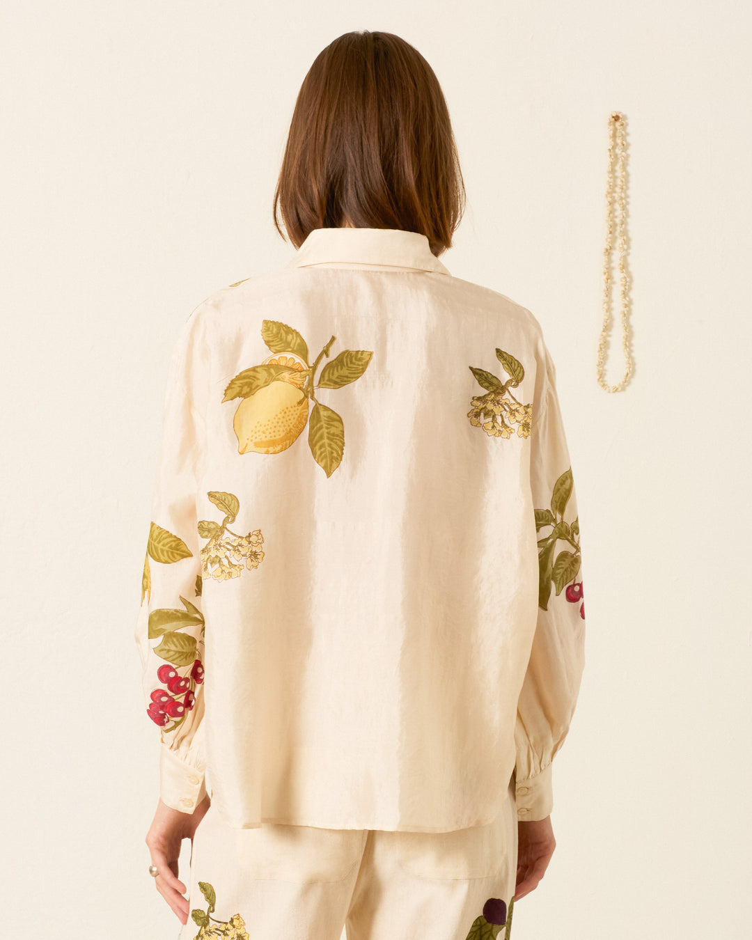 A person stands facing away, wearing the Emery Four Fruit Silk Shirt—a cream-colored, artisan piece with hand-block-printed fruit and leaf motifs. A beaded necklace hangs on the wall to the right.