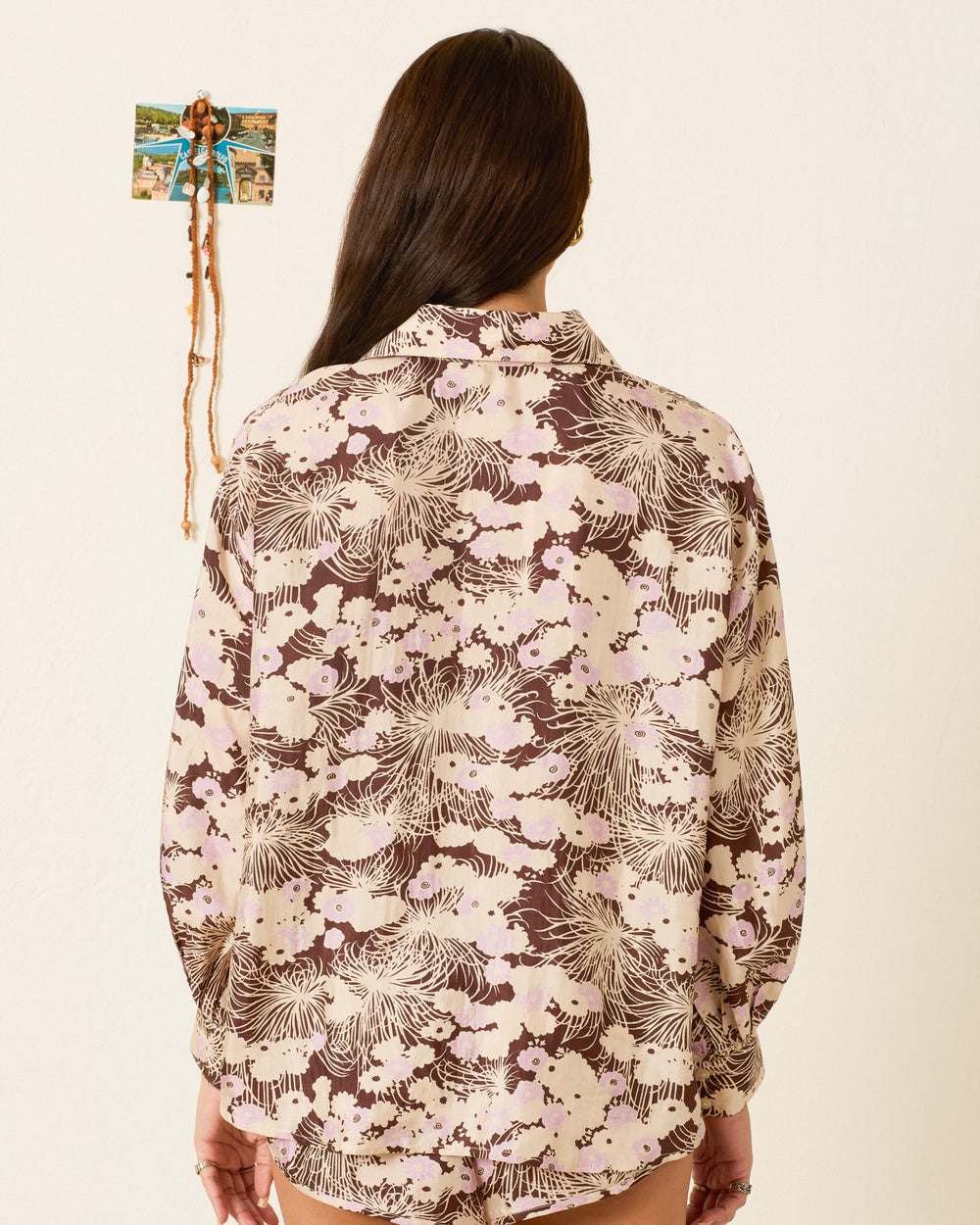 A person with long brown hair, seen from behind, wears the Emery Dandelion Silk Shirt—a loose-fitting, vintage-inspired blouse—while standing before a cream wall decorated with a small photo collage.