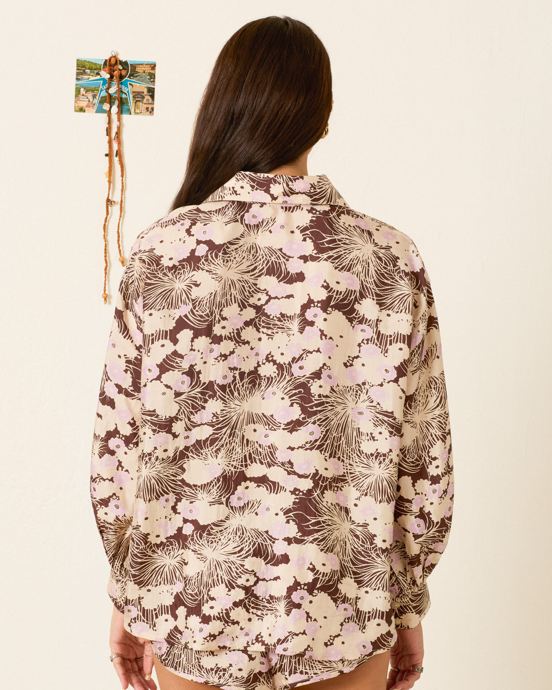 A person with long brown hair, seen from behind, wears the Emery Dandelion Silk Shirt—a loose-fitting, vintage-inspired blouse—while standing before a cream wall decorated with a small photo collage.