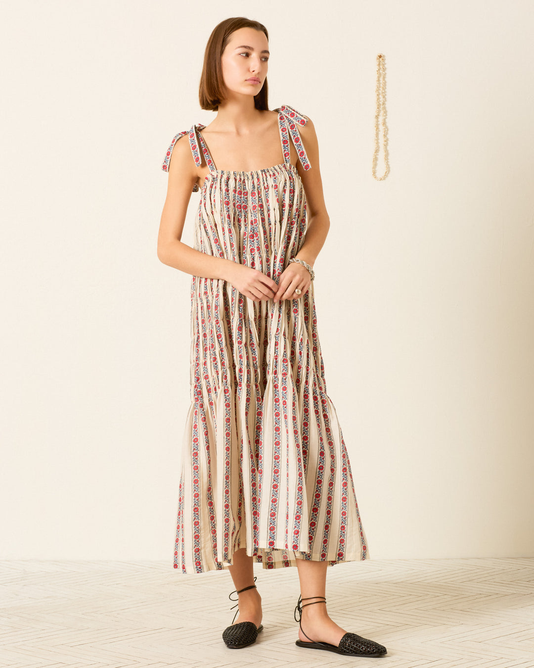 A woman stands indoors against a plain background wearing the Emrys Roseband Stripe Dress, a patterned sleeveless cotton-silk maxi with tie straps, paired with black flats; a necklace hangs on the wall.