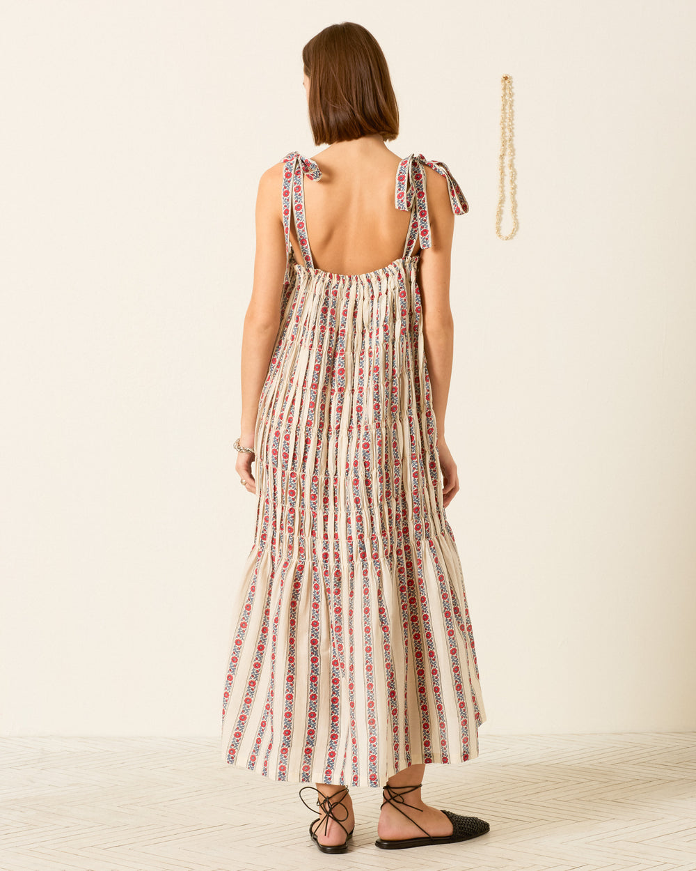 A woman faces away, wearing the Emrys Roseband Stripe Dress with shoulder ties and black lace-up flats, against a plain background featuring a necklace hanging on the wall.