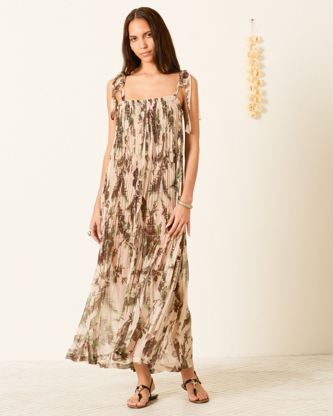 A woman stands against a plain wall, wearing the Emrys Summermoss Dress—a long, sleeveless maxi in cupro-chiffon with a delicate botanical motif and tie straps—paired with sandals. A decorative hanging is visible on the right side of the wall.