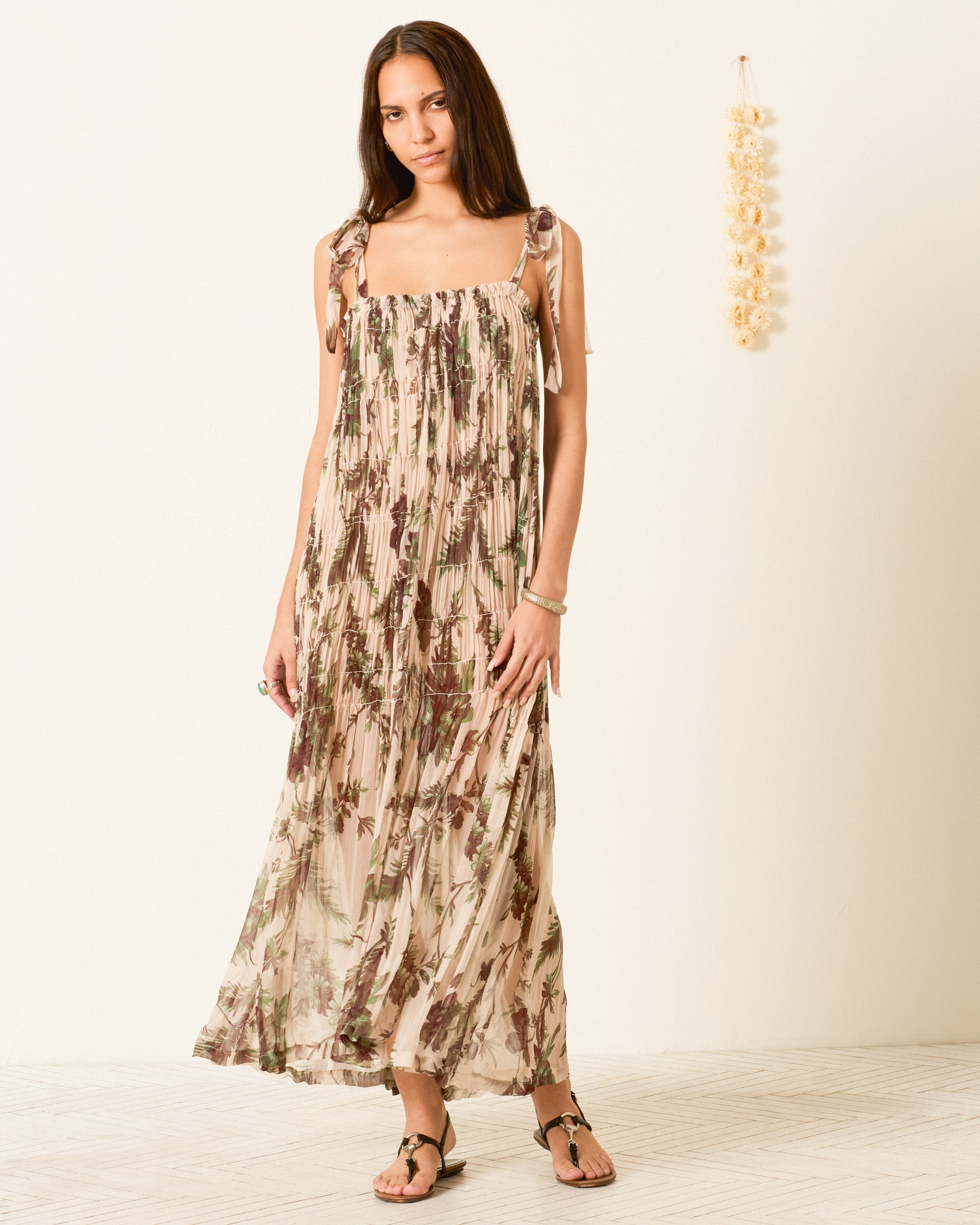 A woman stands against a plain wall, wearing the Emrys Summermoss Dress—a long, sleeveless maxi in cupro-chiffon with a delicate botanical motif and tie straps—paired with sandals. A decorative hanging is visible on the right side of the wall.