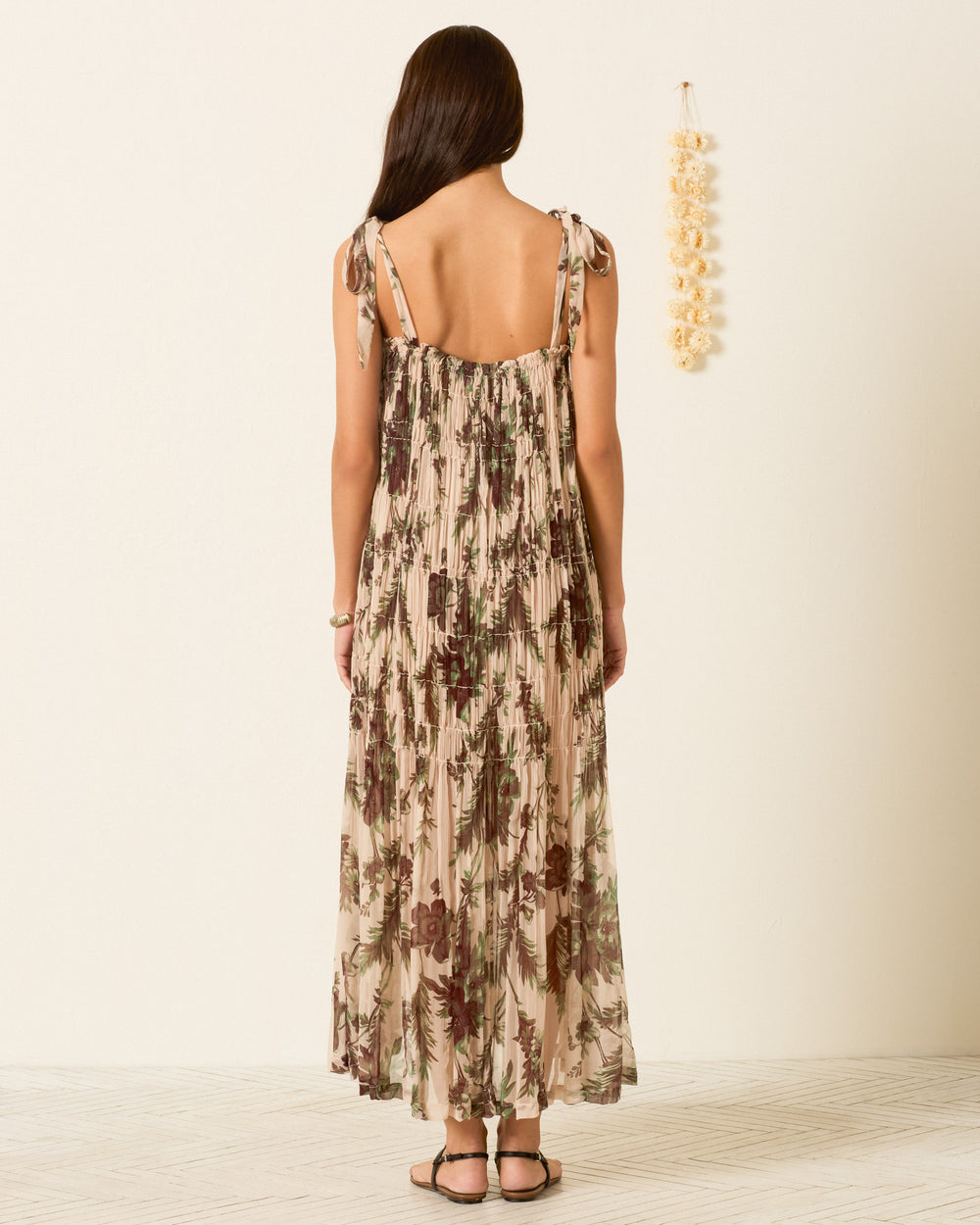 A woman stands indoors, facing away, wearing the sleeveless Emrys Summermoss Dress with a delicate botanical motif and sandals. Decor hangs on the wall to her right, set against a light background and wooden floor.