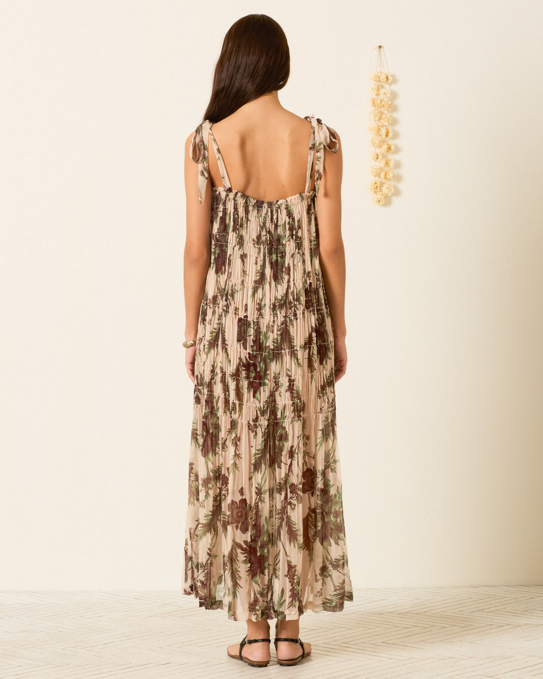 A woman stands indoors, facing away, wearing the sleeveless Emrys Summermoss Dress with a delicate botanical motif and sandals. Decor hangs on the wall to her right, set against a light background and wooden floor.
