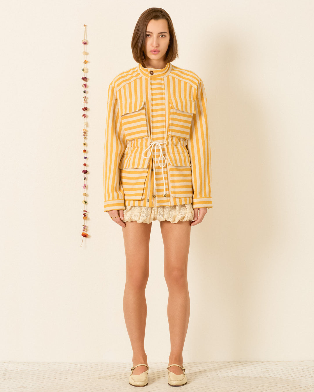 A woman stands against a plain wall wearing the Evelyn Sunlight Stripe Jacket, paired with ruffled shorts and beige shoes. Decorative beads hang vertically to her left.