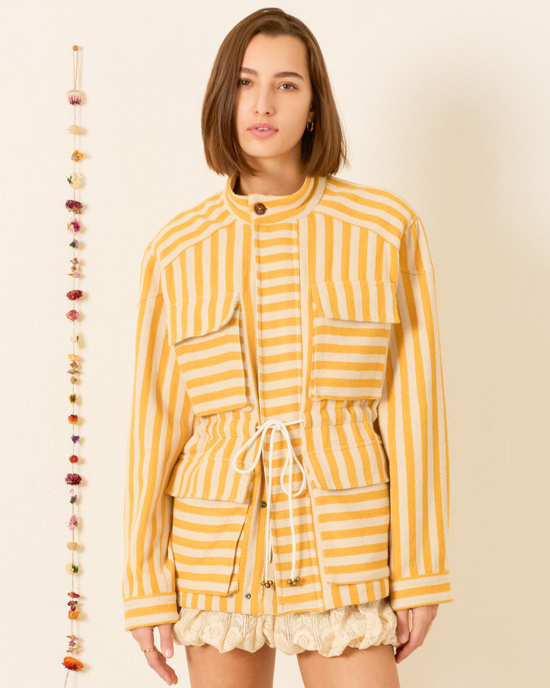 A woman stands indoors beside a hanging string of dried flowers, wearing the Evelyn Sunlight Stripe Jacket—a yellow and white striped utility jacket crafted from soft cotton twill, featuring large pockets and a drawstring waist.