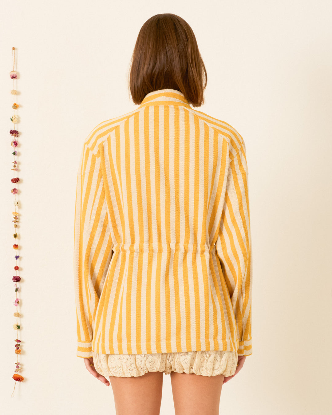 A woman stands facing away, wearing the Evelyn Sunlight Stripe Jacket in yellow awning stripes crafted from cotton twill and cream lace shorts, with a string of dried flowers hanging on the wall to her left.