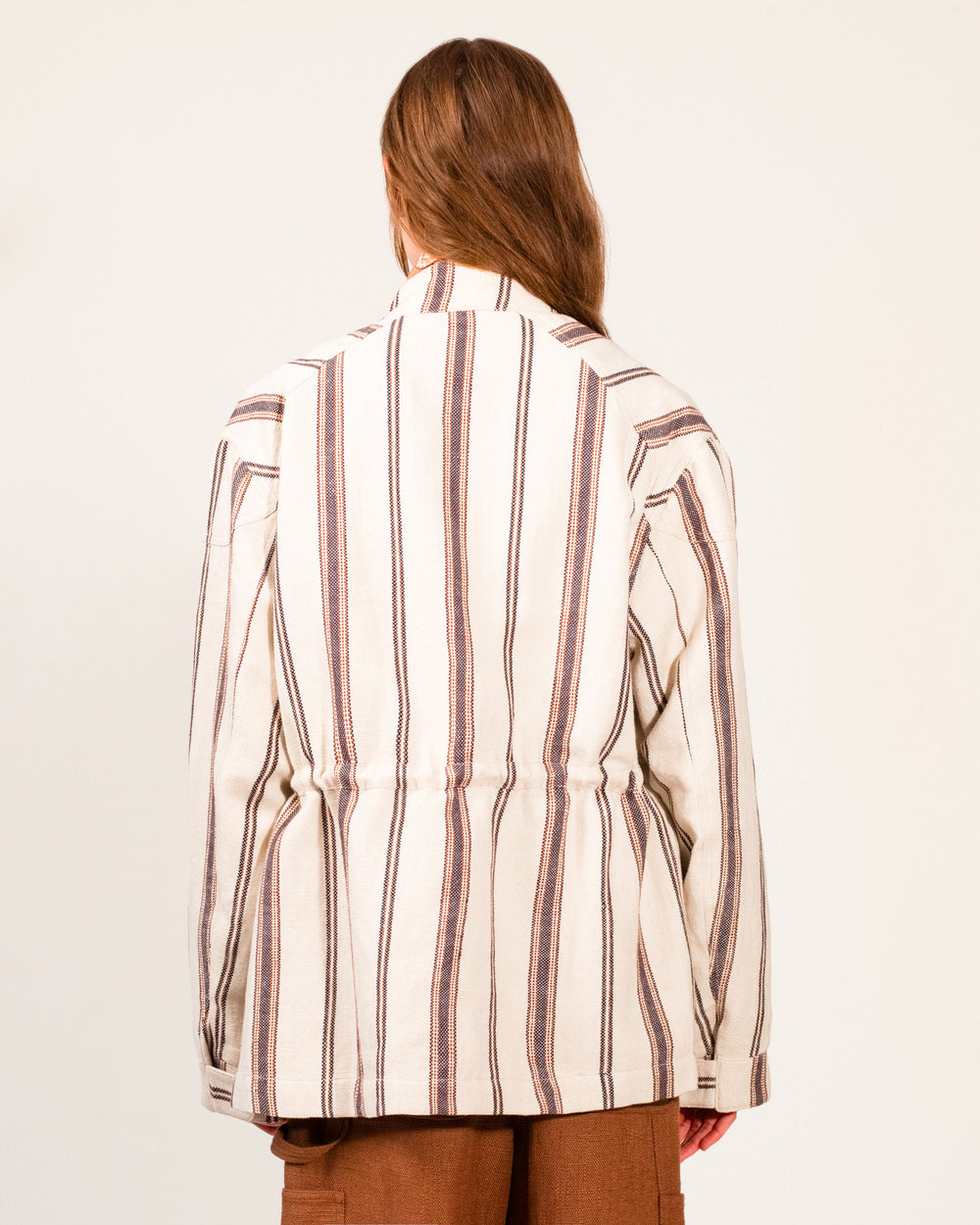 A person with long hair facing away wears the Evelyn Bark Stripe Jacket—a cream linen-cotton utility jacket featuring vertical brown and blue stripes, some hand-embroidered details—paired with brown pants, set against a plain light background.