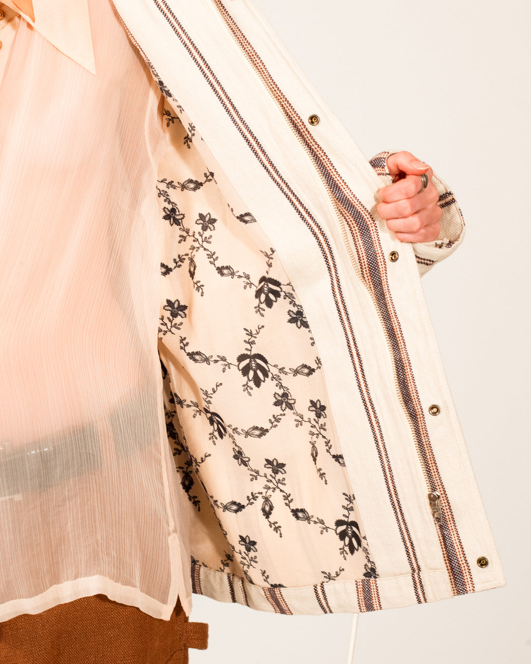 A person opens the Evelyn Bark Stripe Jacket, a light-colored, striped linen-cotton coat, to reveal its floral patterned lining. Only part of their torso and one hand are visible.