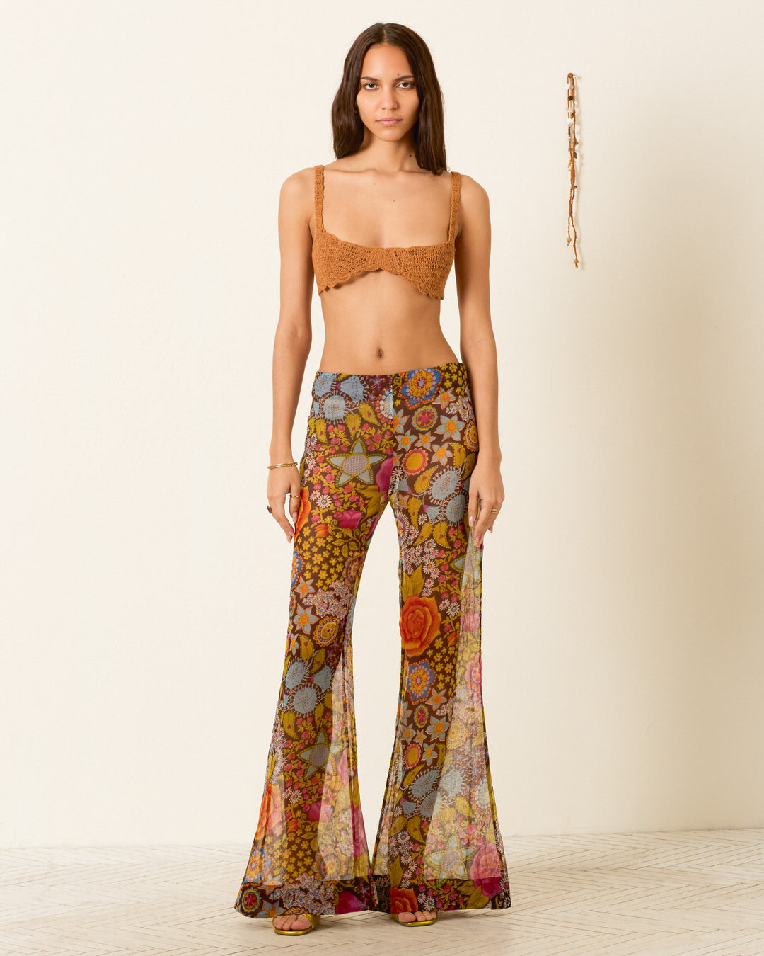 A woman faces forward, wearing a brown crochet bralette and the Farah Bloom Mirage Pant—sheer, high-waisted, with a psychedelic floral motif and bell-bottom flare—against a plain background.