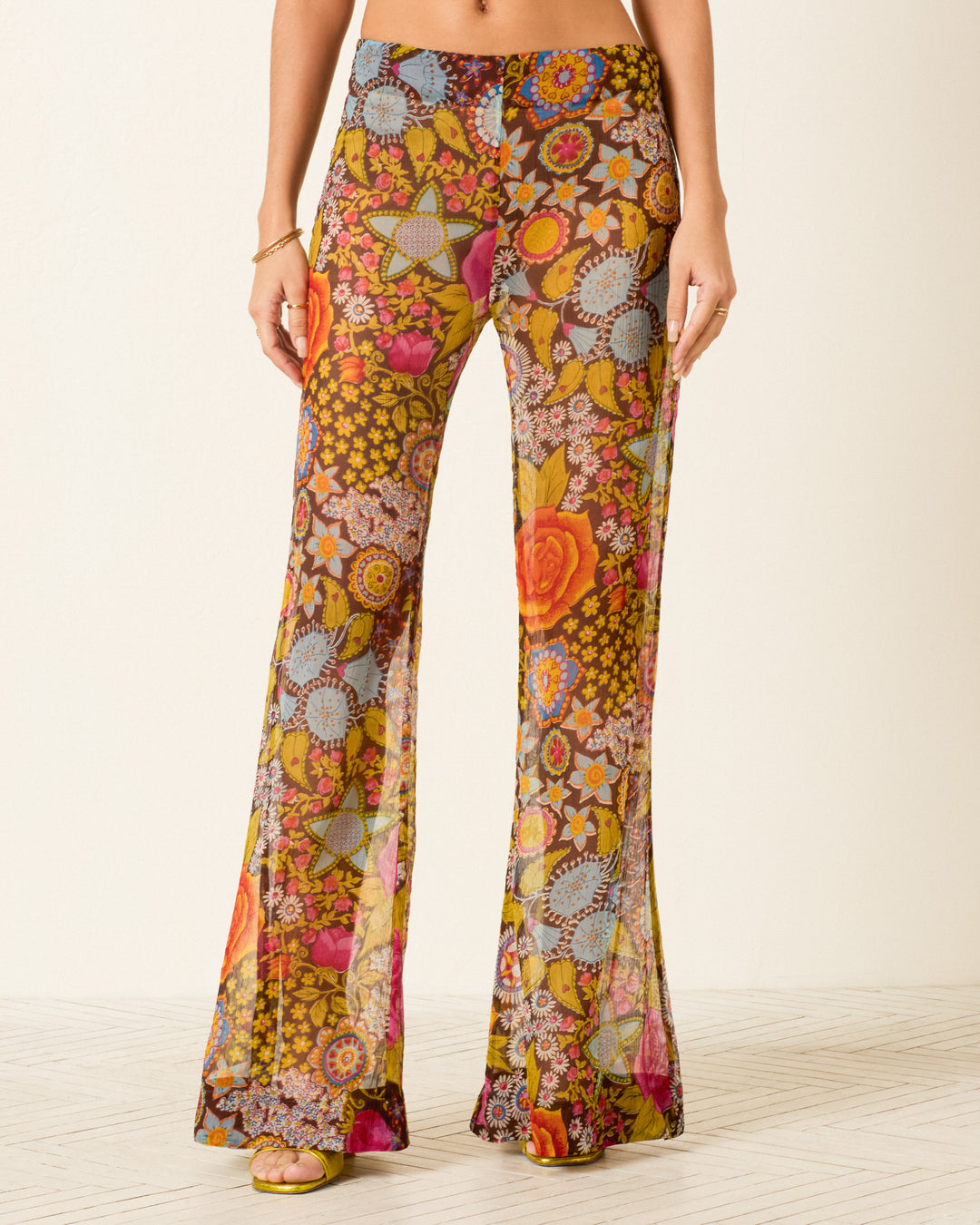 Someone stands indoors on a light-colored floor wearing Farah Bloom Mirage Pant, featuring a sheer, high-waisted design with vibrant geometric and psychedelic floral patterns.