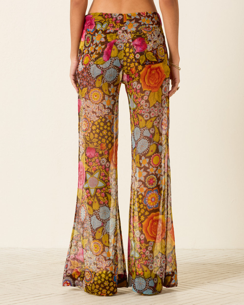 A person wears the Farah Bloom Mirage Pant—sheer, high-waisted pants with psychedelic floral motifs, colorful geometric patterns, and wide bell-bottom flare—photographed from the back.