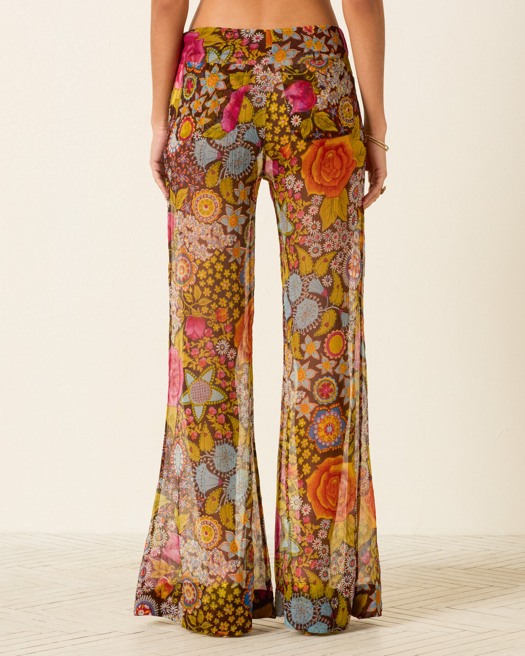 A person wears the Farah Bloom Mirage Pant—sheer, high-waisted pants with psychedelic floral motifs, colorful geometric patterns, and wide bell-bottom flare—photographed from the back.