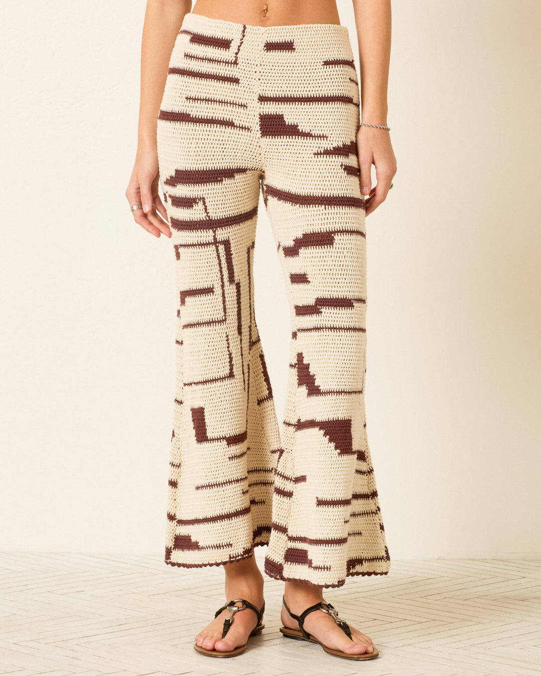 A person wears the Farah Auburn Driftmap Pant—high-waisted, cream-colored hand-crocheted cotton pants with brown, ikat-inspired geometric patterns and bell-bottom flares—paired with black sandals and standing against a light background.