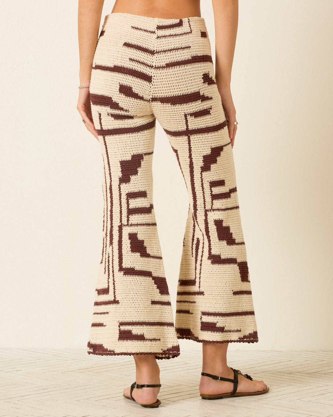 Wearing the Farah Auburn Driftmap Pant—cream and brown, hand-crocheted cotton pants featuring a geometric ikat-inspired pattern and bell-bottom flare—with black sandals, the person is shown from the back standing indoors on a light floor.