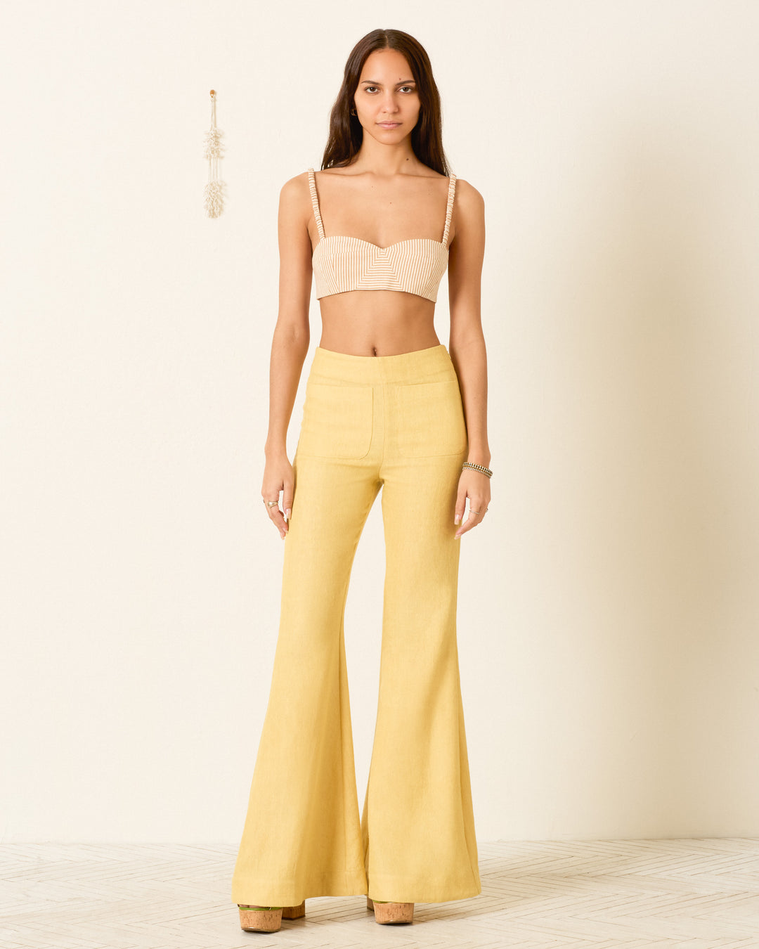 A woman stands against a plain wall, wearing a beige cropped top and the Farah Sunlight Linen Pant: high-waisted yellow pants featuring a dramatic bell-bottom flare crafted from linen and wool, paired with platform shoes.