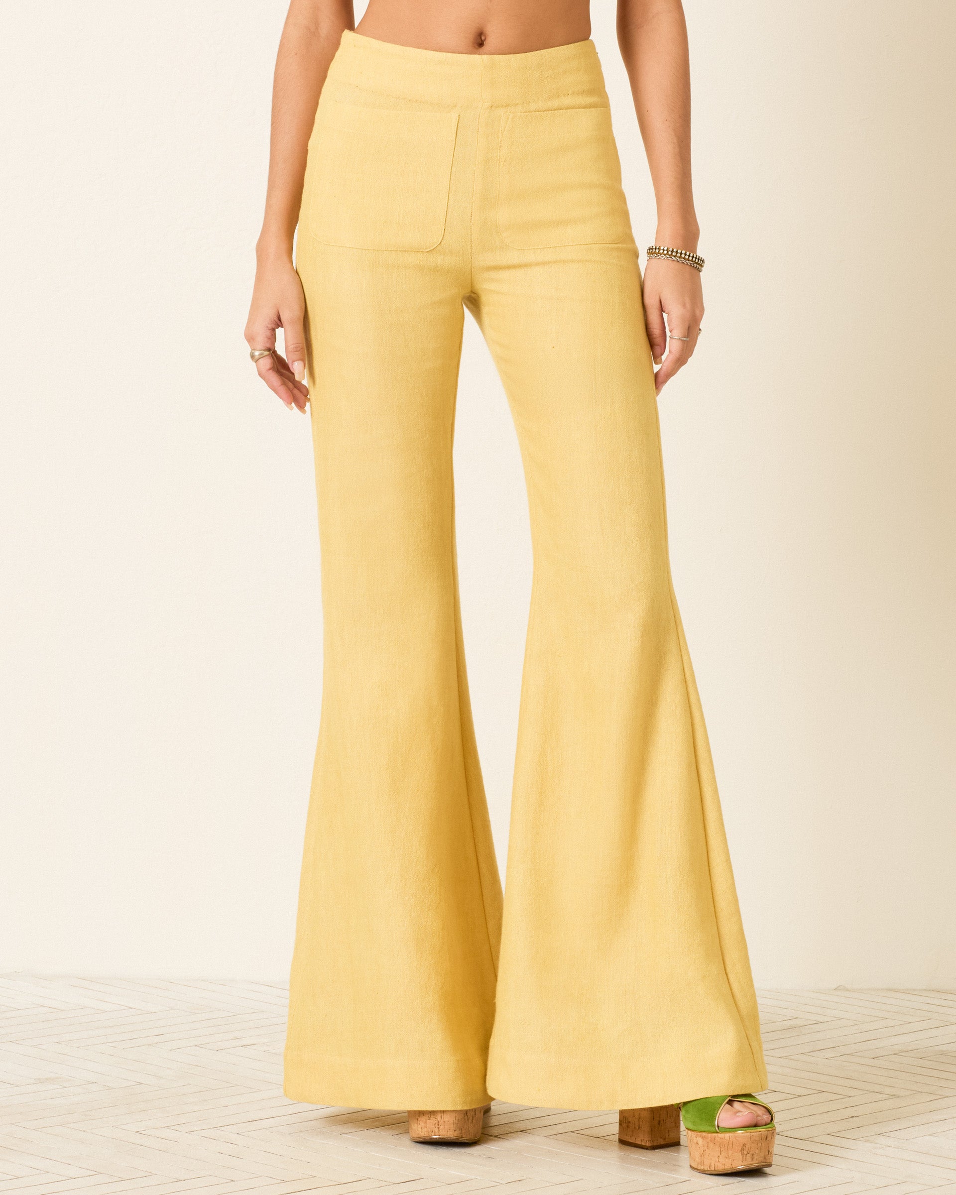 A person wearing the Farah Sunlight Linen Pant—a high-waisted, yellow bell-bottom flare pant crafted from a blend of linen and wool—paired with platform sandals and standing against a light background.