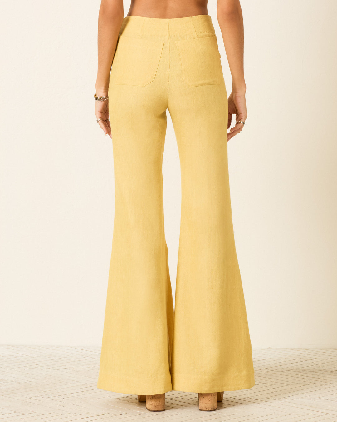 Person wearing Farah Sunlight Linen Pant—high-waisted, yellow linen and wool bell-bottom flare pants with back pockets—standing barefoot on a light-colored floor, shown from the back.