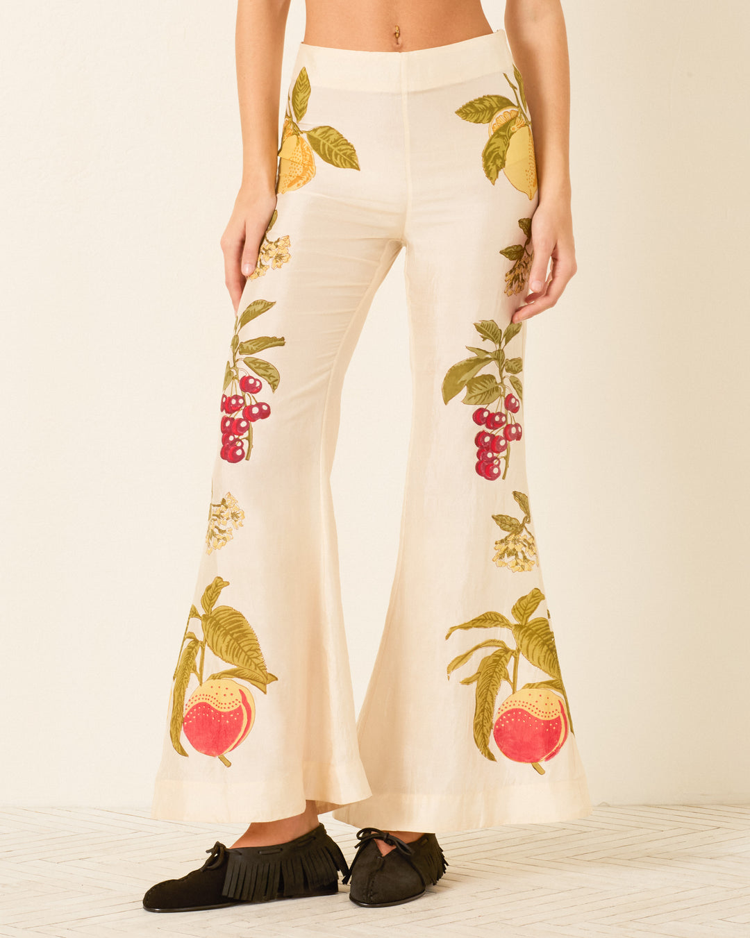 A person stands against a light background wearing the Farah Four Fruit Silk Pant—cream-colored, high-waisted pants adorned with fruit and floral prints—and pairs them with black fringe shoes.