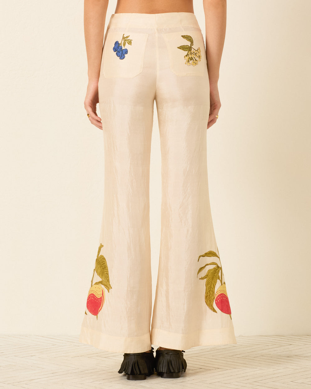 A person is seen from the back wearing the Farah Four Fruit Silk Pant—cream-colored, high-waisted pants adorned with embroidered fruit and plant motifs and finished with a subtle bell-bottom flare—while standing on a light-colored floor.