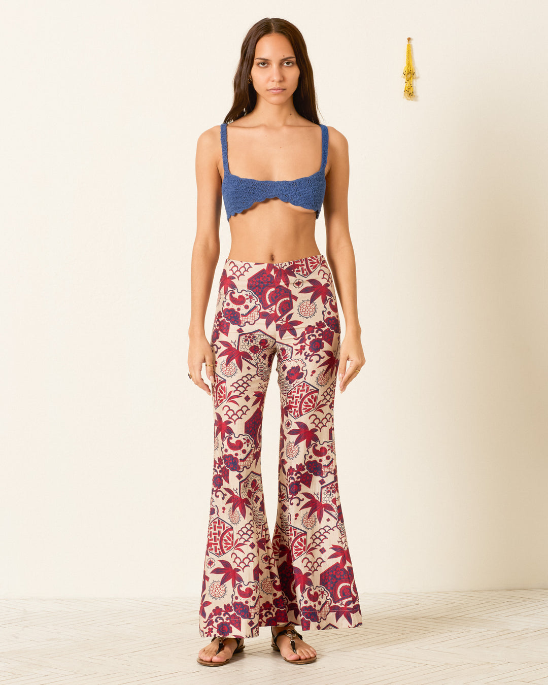 A woman stands against a plain wall wearing a blue bralette, black sandals, and the Farah Crimson Hive Silk Pant featuring vintage wagara prints and a high-waisted bell-bottom flare design.