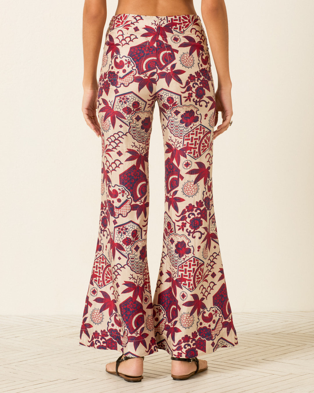 Person wearing Farah Crimson Hive Silk Pant—high-waisted, wide-leg with a red and beige floral and geometric pattern, bell-bottom flare style—viewed from the back while standing on a light patterned floor.