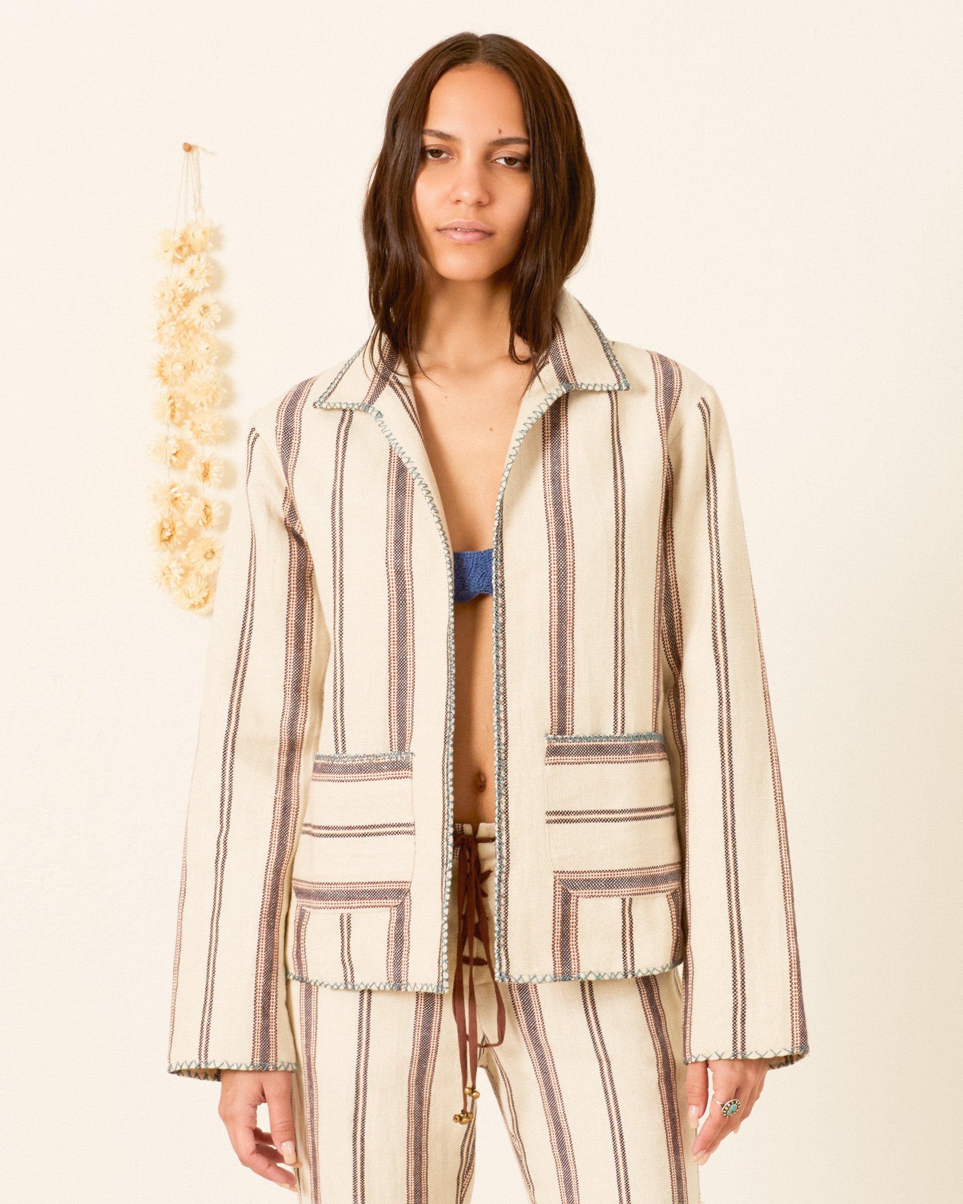 A person wears the Francis Bark Stripe Jacket, made from linen and cotton, with matching pants, standing before a plain wall decorated with a hanging string of dried flowers.