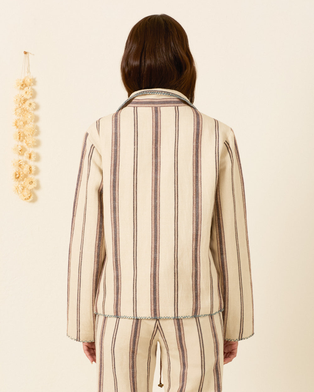 A person stands facing away, wearing the Francis Bark Stripe Jacket— a cream woven linen-cotton piece— beside a hanging garland on a light wall.