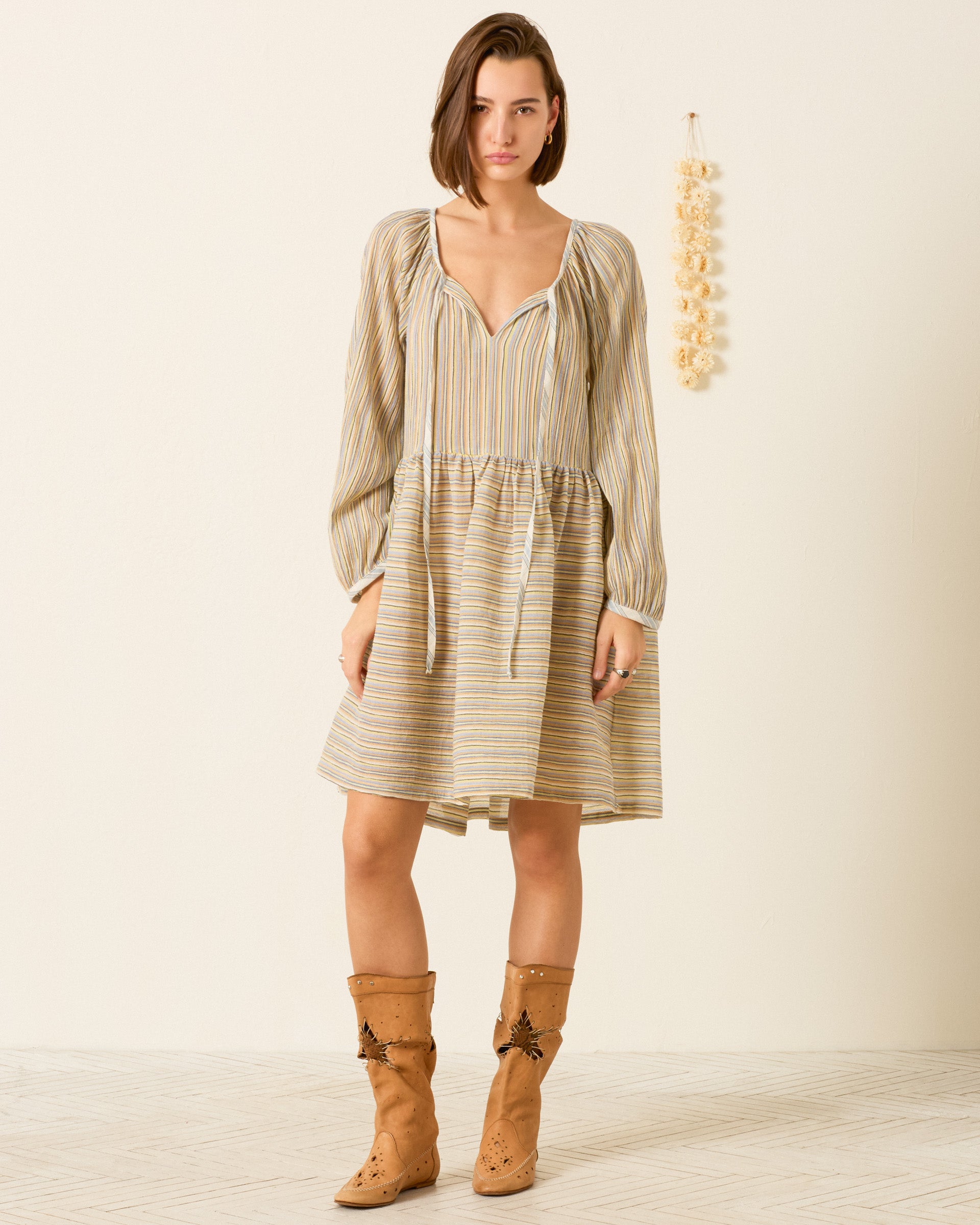 A woman stands indoors wearing the Harriet Sandlilt Stripe Dress—relaxed, yarn-dyed cotton with stripes—paired with tan knee-high boots; a decorative hanging is visible on the wall behind her.