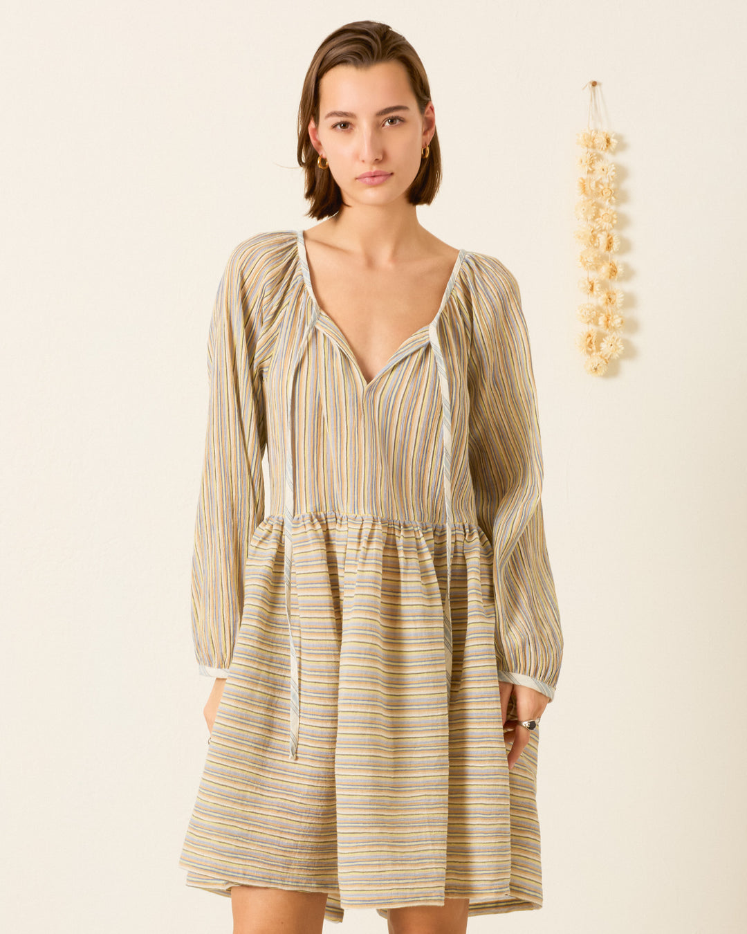A woman models the Harriet Sandlilt Stripe Dress—a loose, long-sleeved, yarn-dyed cotton dress with a V-neckline—indoors beside a hanging decoration; this stylish piece is perfect as a cozy beach cover-up.
