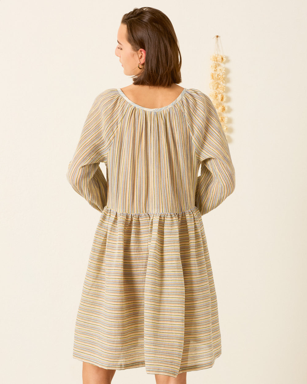 A woman faces away, wearing the Harriet Sandlilt Stripe Dress—a yarn-dyed cotton, knee-length dress with yellow, blue, and beige vertical stripes. A small hanging decoration is on the wall next to her.
