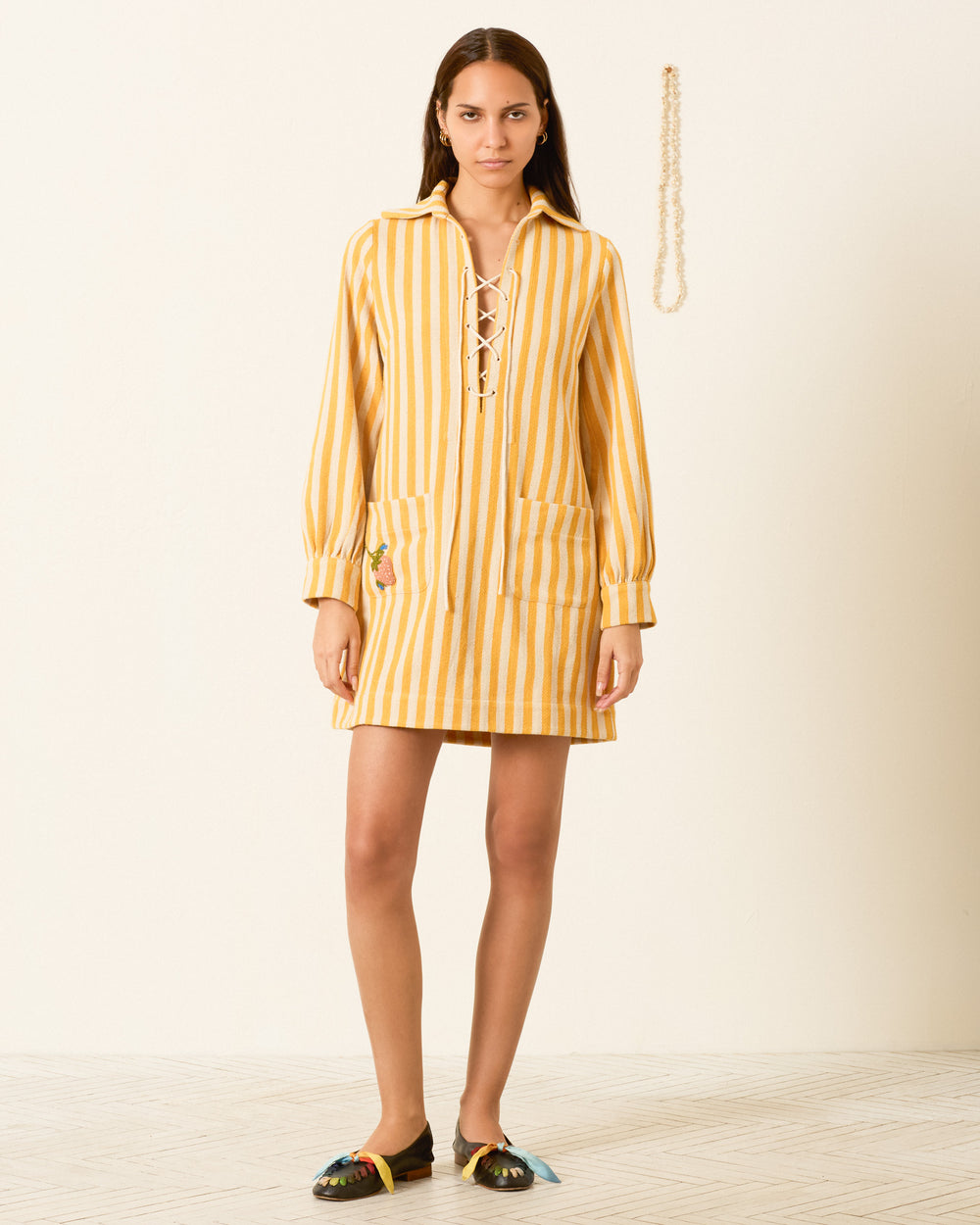 A woman stands against a plain wall wearing the Isadora Sunlight Stripe Dress, featuring yellow and white stripes, cotton twill fabric, front pockets, and a lace-up neckline, paired with flat shoes.