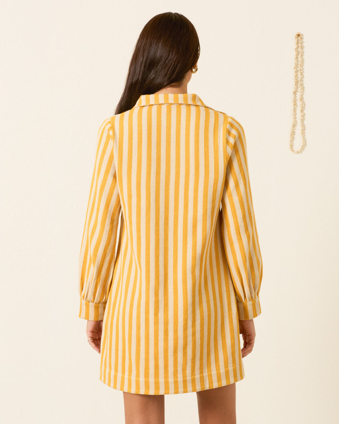 A woman with long brown hair models the Isadora Sunlight Stripe Dress, featuring yellow and white vertical stripes in cotton twill, viewed from the back. A single necklace is displayed on the wall to her right.