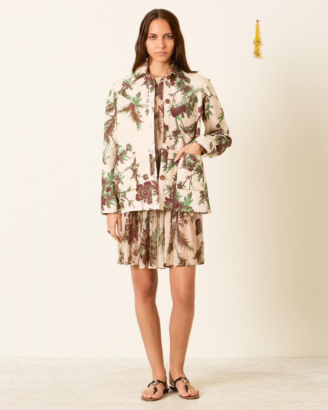 A woman stands against a plain wall, wearing the vintage-inspired Jude Summermoss Jacket and skirt adorned with a botanical motif, paired with black sandals.