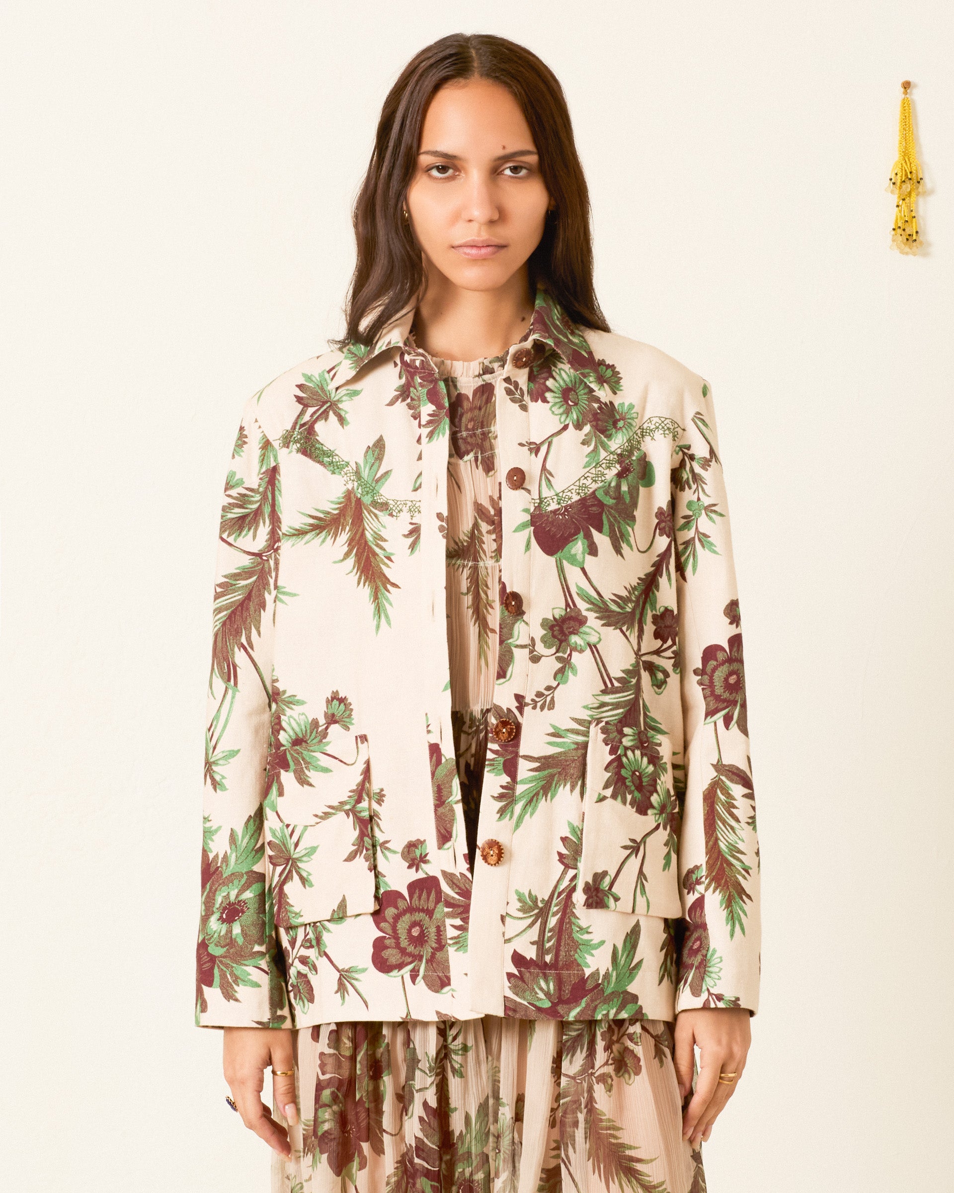 A woman with long brown hair stands against a plain light background, wearing the vintage-inspired Jude Summermoss Jacket in beige Khadi cotton with green and brown botanical motifs, layered over a matching dress.