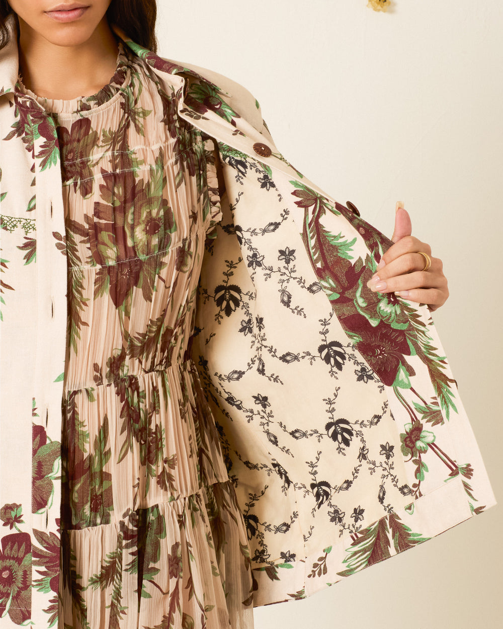 A woman holds open the vintage-inspired Jude Summermoss Jacket, crafted from Khadi cotton with a beige floral-patterned exterior, to reveal its inner lining adorned with a contrasting botanical motif. She pairs it with a pleated dress in complementary tones.