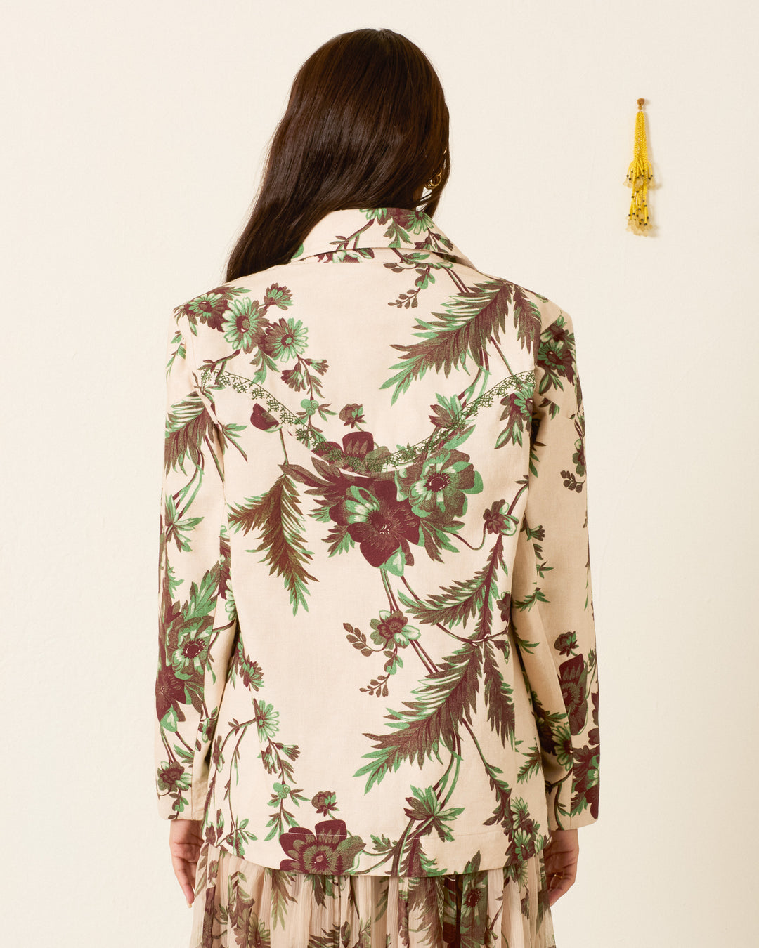 A person with long brown hair, standing with their back to the camera against a plain wall, wears the Jude Summermoss Jacket—a vintage-inspired cream Khadi cotton piece featuring green and maroon botanical motifs.