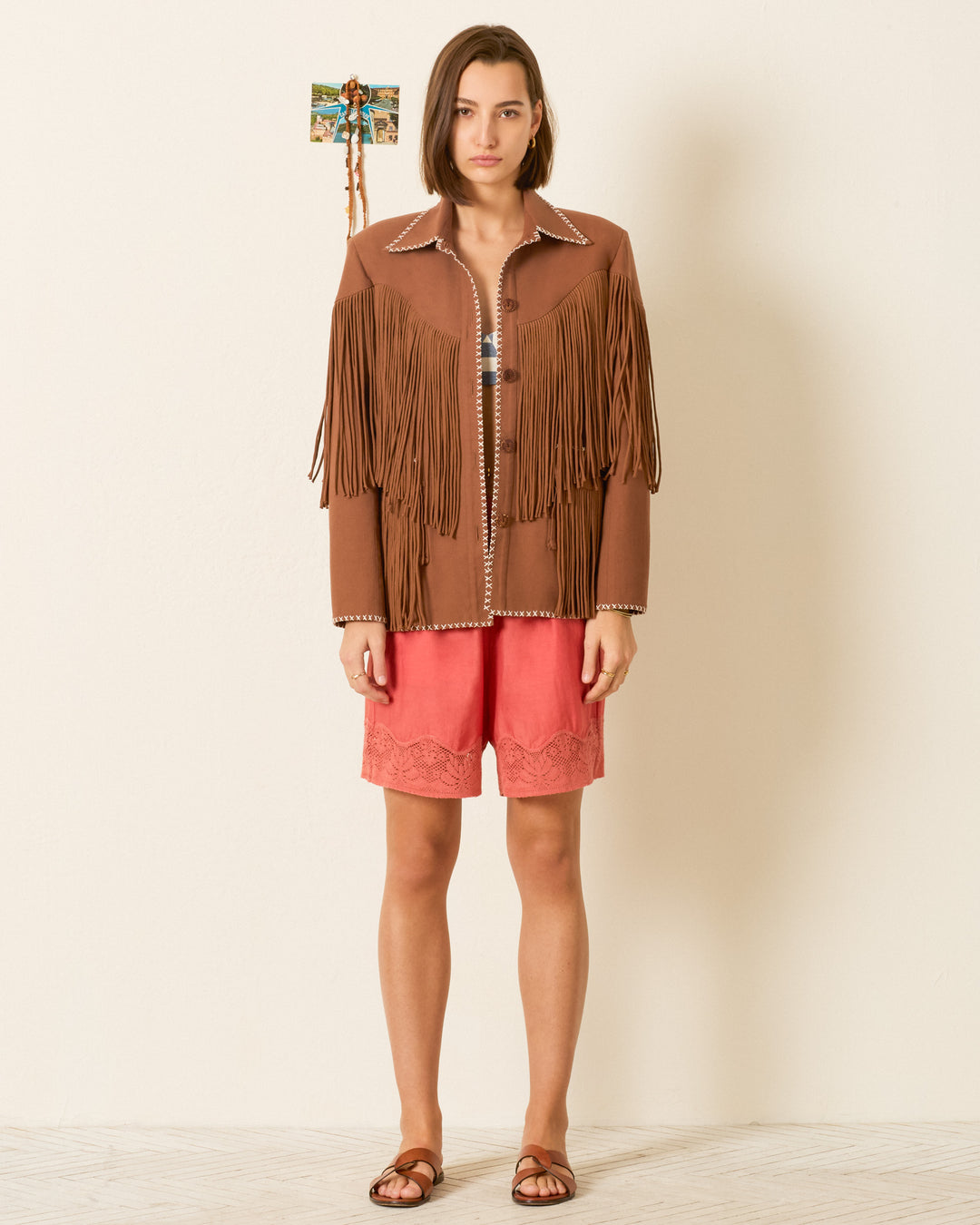 A person stands against a plain wall wearing the Jude Clove Fringe Jacket, paired with coral shorts with lace trim and brown sandals.