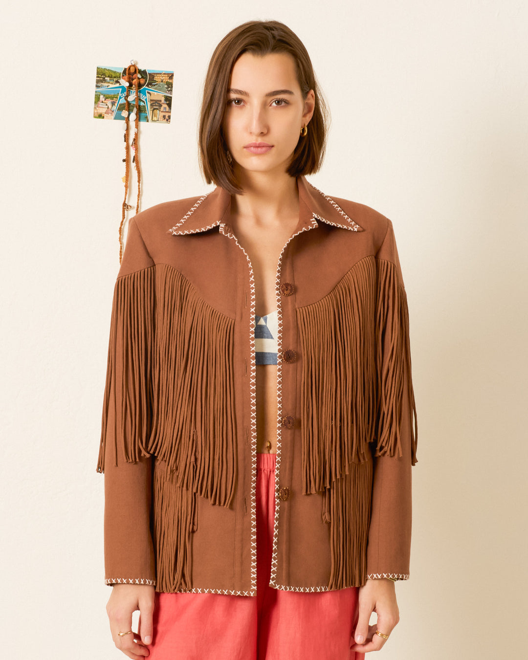 A woman stands against a plain wall, decorated with a small photo and ribbon, wearing the Jude Clove Fringe Jacket and red pants.