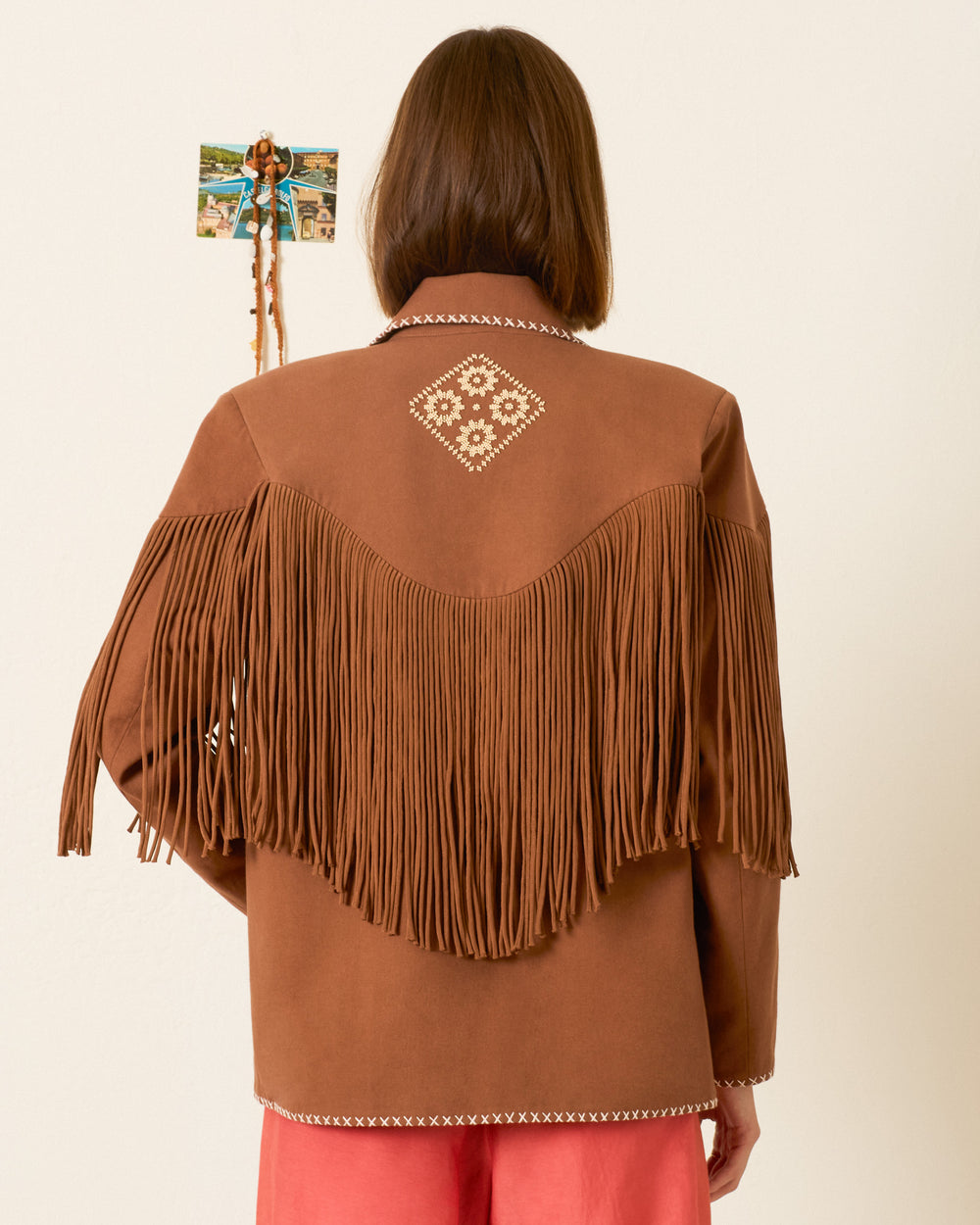 A person with shoulder-length brown hair wears the Jude Clove Fringe Jacket, featuring long fringing and white embroidery in a Western-inspired style, standing with their back to the camera. A small decoration is pinned to the wall.