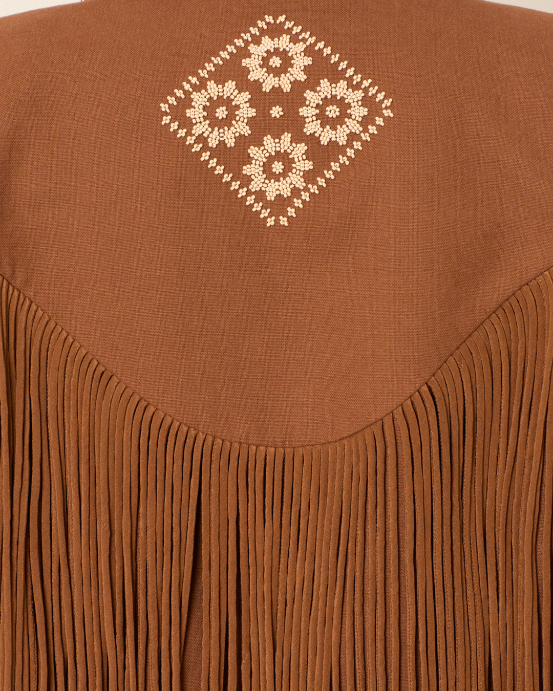 The Jude Clove Fringe Jacket features brown fabric with long vertical fringe and a white embroidered diamond floral pattern centered near the top, evoking a Western-inspired style.