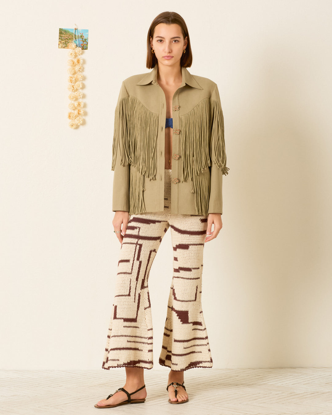 A person stands indoors wearing the Jude Moss Fringe Jacket, styled with patterned wide-leg pants and black sandals, with a small wall decoration visible in the background.