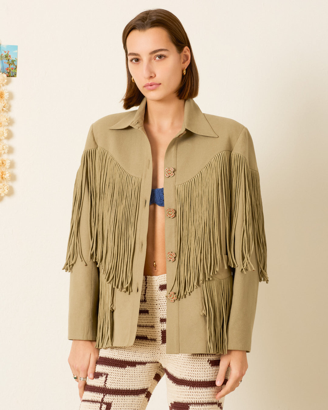Wearing the Jude Moss Fringe Jacket, a woman stands against a plain background, layering it over a blue bralette and patterned knit pants.