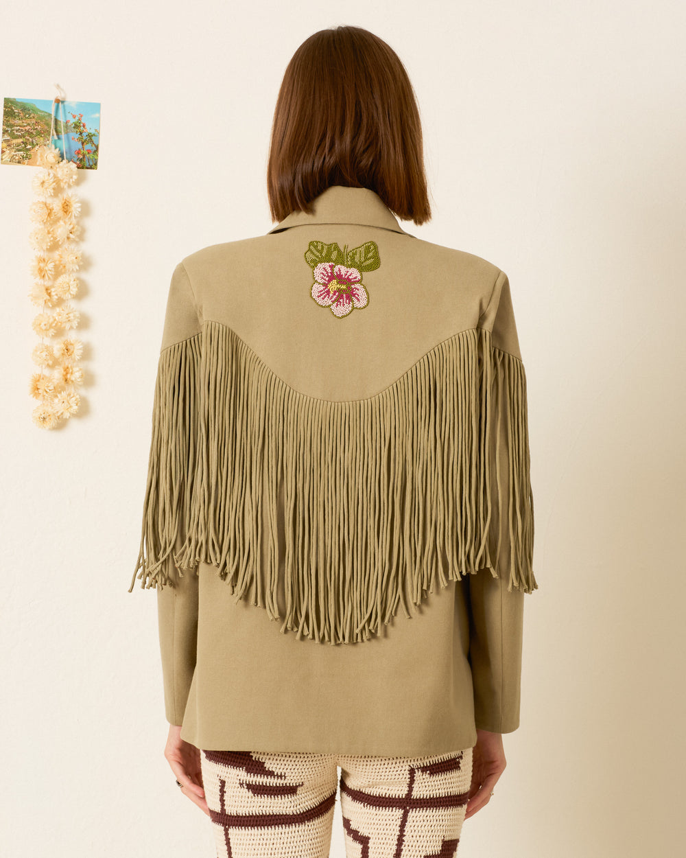 A person stands facing away, wearing the Jude Moss Fringe Jacket—a Western-inspired khaki piece with a floral patch on the back—paired with patterned knit pants. The textured cotton of the jacket lends unique dimension, complemented by a photo, lei, and shell decor hanging on the wall to the left.