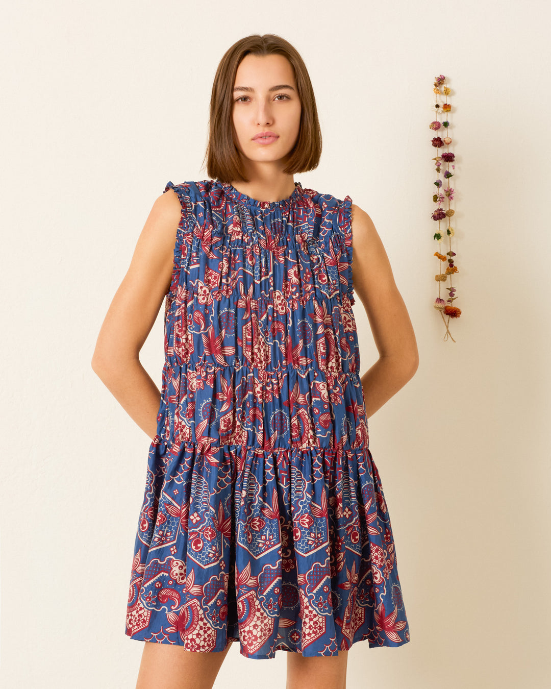 A woman stands against a plain wall with a small hanging of dried flowers on her right, wearing the Jule Delft Hive Silk Dress— a sleeveless, loose-fitting dress in blue and red with a subtle wagara print.