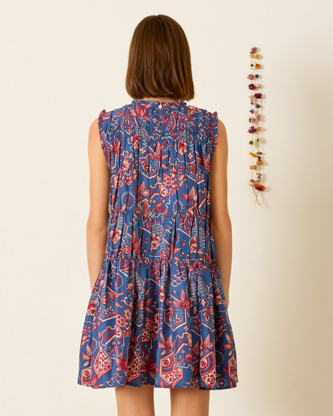 A woman stands facing away, wearing the Jule Delft Hive Silk Dress—a blue sleeveless dress adorned with a red and white floral pattern inspired by traditional wagara prints. Dried flowers are hung on the wall to her right.