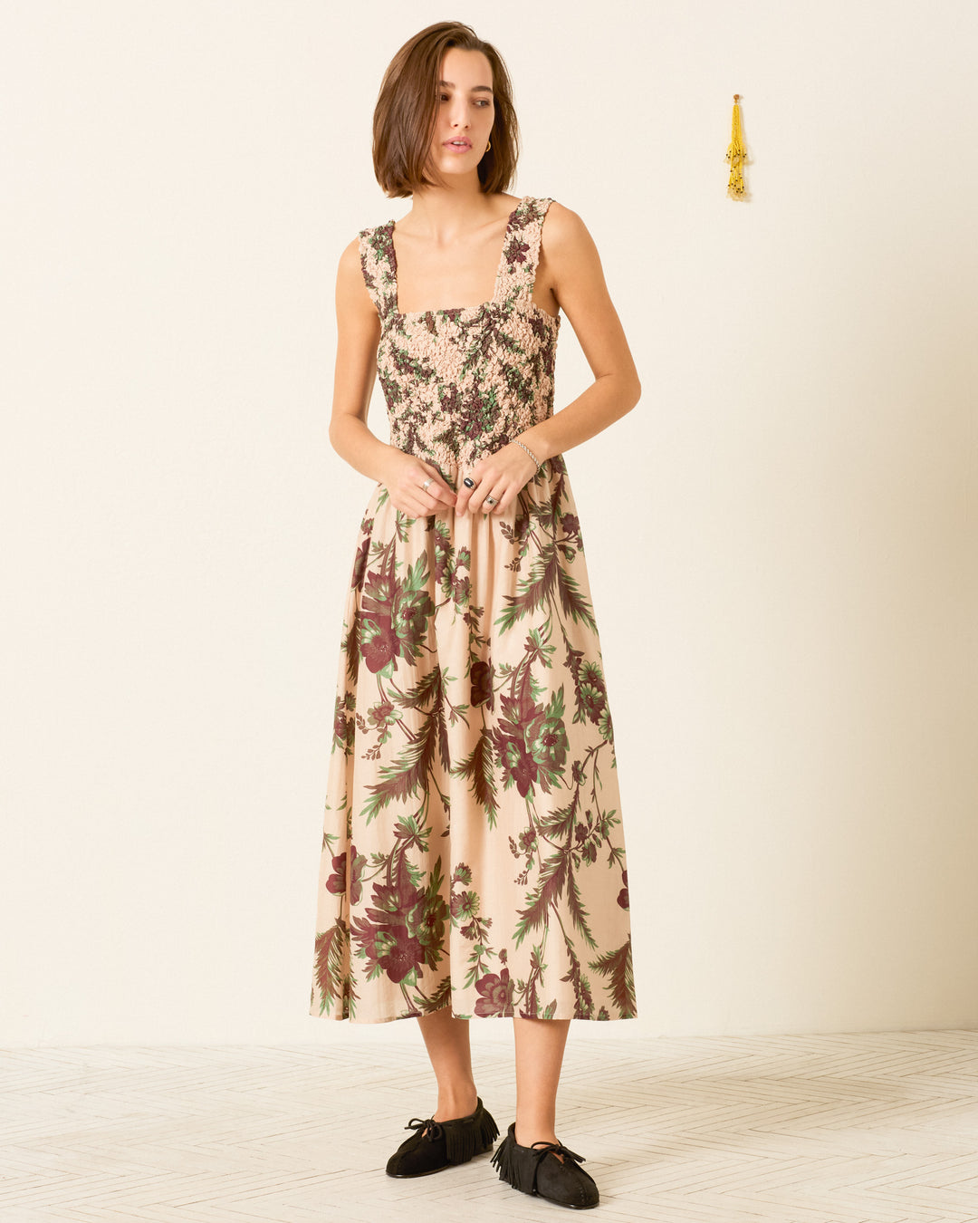 A woman wearing the Kay Summermoss Dress—a sleeveless cotton-silk midi dress with a botanical motif, smocked bodice, and flowing skirt—stands against a plain off-white wall, paired with black flats.