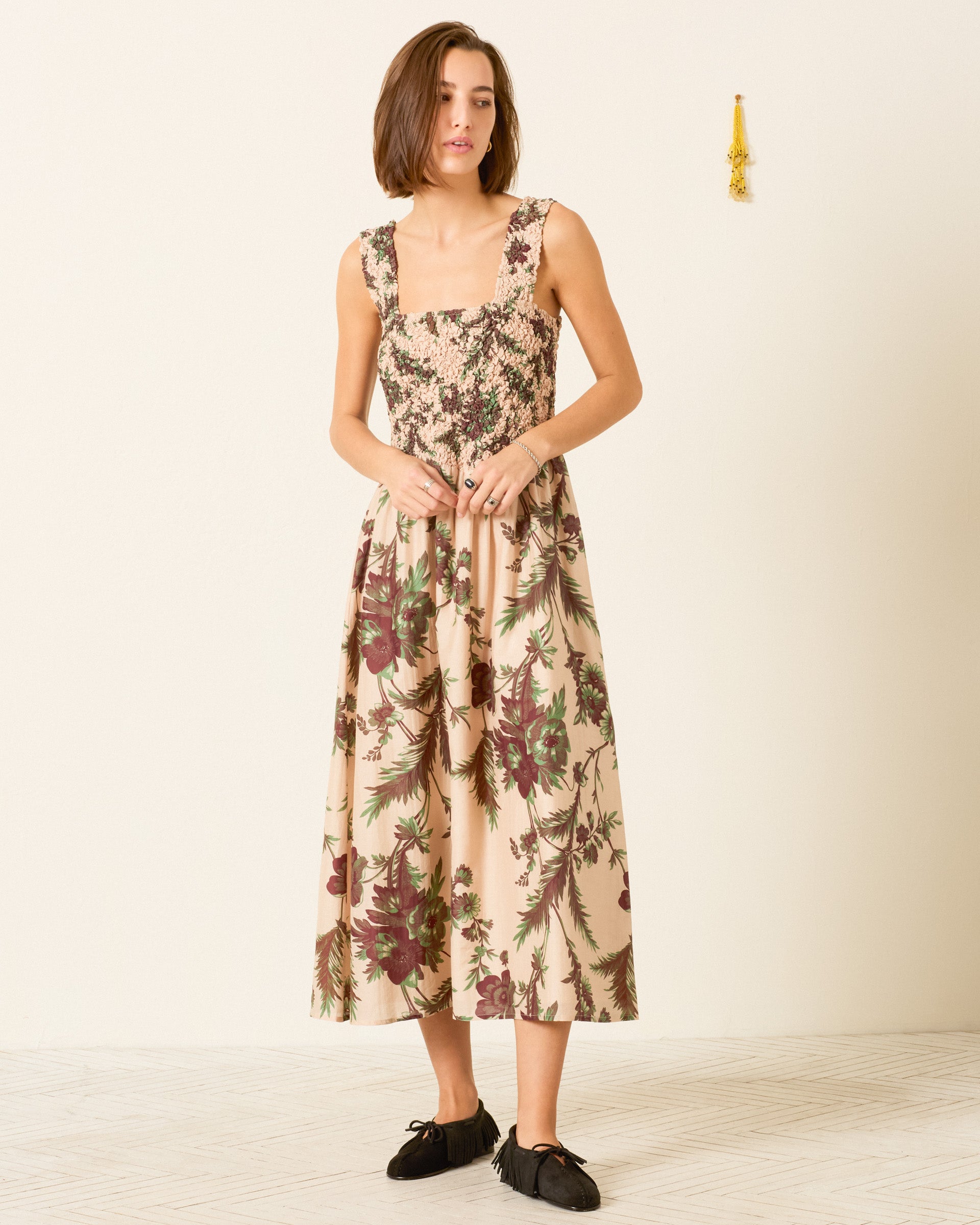 A woman wearing the Kay Summermoss Dress—a sleeveless cotton-silk midi dress with a botanical motif, smocked bodice, and flowing skirt—stands against a plain off-white wall, paired with black flats.